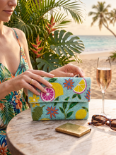 Load image into Gallery viewer, A woman at a beach table holds a turquoise clutch bag with a colorful tropical lemon and flower print. A champagne flute, sunglasses, and a small gold case sit on the table, with palm trees and sand in the background.