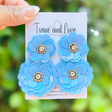Load image into Gallery viewer, Blue floral earrings on a card held by a hand with a green leafy background