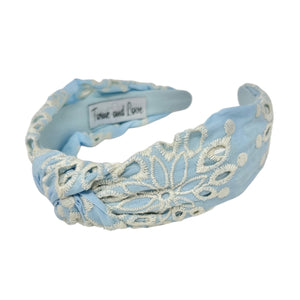 Light blue headband with intricate white floral embroidery, displayed in a side view. The soft texture and elegant design convey a stylish, timeless appeal.