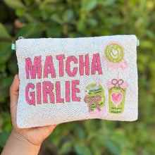 Load image into Gallery viewer, A hand holds a light green beaded pouch with pink 'MATCHA GIRLIE' text and colorful matcha-themed illustrations, set against a blurred green foliage background.