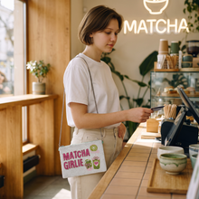 Load image into Gallery viewer, A woman in a white t-shirt and beige pants stands at a cafe counter, holding a card near a white shoulder bag with pink and green graphics that read 'MATCHA GIRLIE'. The background shows a warm wood interior, a glowing 'MATCHA' sign, and cafe items.
