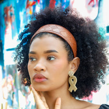Load image into Gallery viewer, Curly-haired model showcasing a wide cognac brown headband and sparkling gold ribbon-shaped earrings against a colorful artistic backdrop.