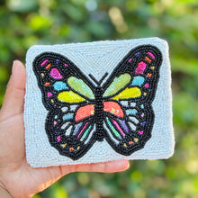 Load image into Gallery viewer, A hand holds a square, white beaded pouch featuring a colorful embroidered butterfly design on the front, set against a blurred green outdoor background.