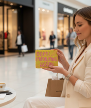 Load image into Gallery viewer, A woman in a cream blazer sits in a shopping mall holding a yellow pouch with pink text that reads " I'M BUYING AGAIN". A Malone shopping bag and a coffee cup are visible on a table in the foreground, with blurred mall storefronts in the background.
