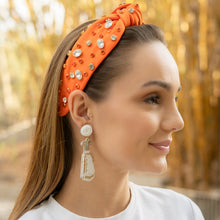 Load image into Gallery viewer, Woman wearing an orange headband with decorative elements and earrings, set against a blurred natural background.