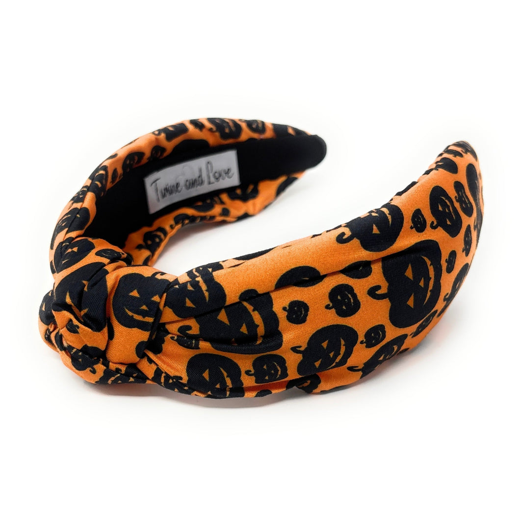 Orange knotted headband with black Halloween-themed prints, featuring pumpkins and cats. The design is playful, capturing a festive Halloween vibe.