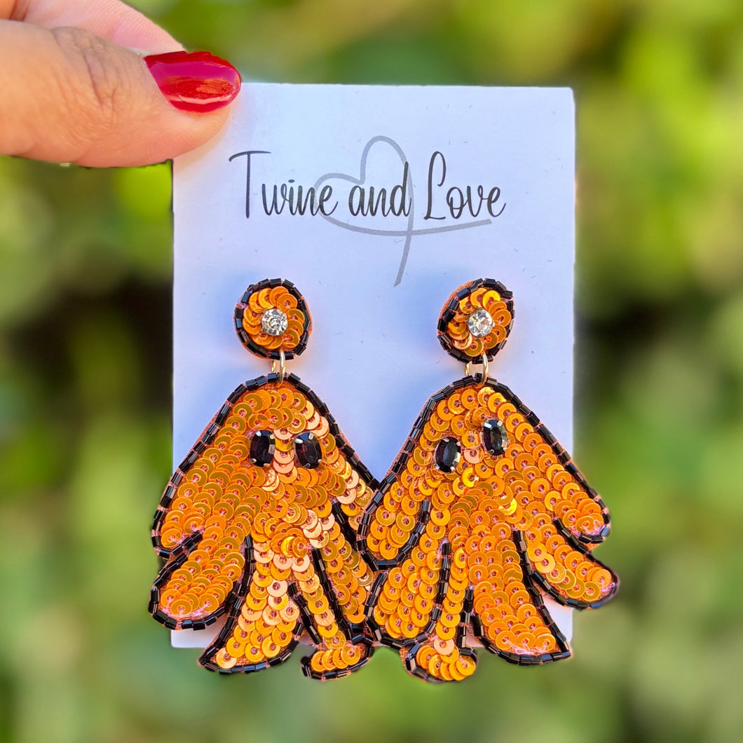 Orange ghost earrings decorated with sequins and black seed beads on a white background. The playful design conveys a fun, festive, and whimsical Halloween theme.