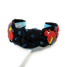 Load image into Gallery viewer, A black fabric headband with a twisted knot center, decorated with colorful floral beadwork in red, yellow, and blue, displayed on a white stand against a white background.