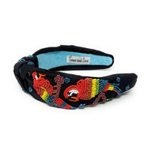 Load image into Gallery viewer, A black fabric headband with a knotted center, decorated with colorful beaded parrots designs in red, yellow, green, and brown, and small blue beaded flowers. The inside of the headband features a light blue felt lining with a white label that reads 'Twine and Love'.