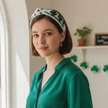 Load image into Gallery viewer, Woman in a green shirt and white pants holding a mug, wearing a shamrock headband, in a room with decorative elements.