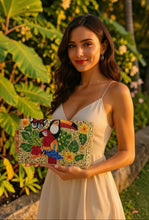 Load image into Gallery viewer, A woman in a cream-colored spaghetti-strap dress holds a rectangular clutch bag decorated with colorful embroidered birds and tropical leaves, set against a lush tropical garden background.