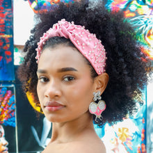 Load image into Gallery viewer, Woman wearing a pink pearl-embellished knotted headband and pink tennis racket statement earrings, styled with a black off-the-shoulder dress in front of colorful artwork.