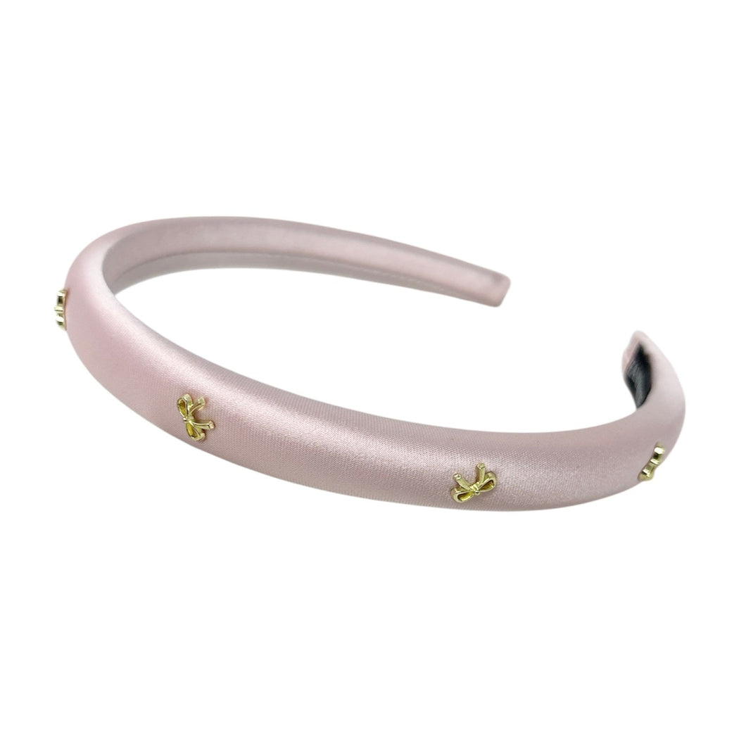 A satin pink headband adorned with small gold embellishments lies on a white background. The delicate design conveys elegance and sophistication.