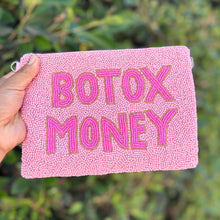 Load image into Gallery viewer, A hand holds a pink beaded pouch with the words 'BOTOX MONEY' in raised pink letters outlined in gold, set against a blurred green foliage background.