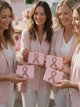 Load image into Gallery viewer, Four women in matching light pink blazers and white tops smile while holding small pink pouches with a large pink awareness ribbon on the front, set in a softly lit pink-themed indoor setting.