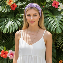 Load image into Gallery viewer, A woman wearing a light purple knotted headband and a white spaghetti-strap dress stands in front of a lush tropical background of green leaves and colorful hibiscus flowers.