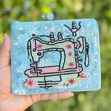 Load image into Gallery viewer, Hand holding a blue embroidered wallet with a sewing machine design against a blurred green background