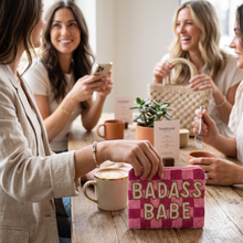 Load image into Gallery viewer, Four women gathered around a wooden table in a bright cafe or studio setting, smiling and laughing. In the foreground, a woman in a beige blazer holds a pink woven clutch with the words 'BADASS BABE' in gold.
