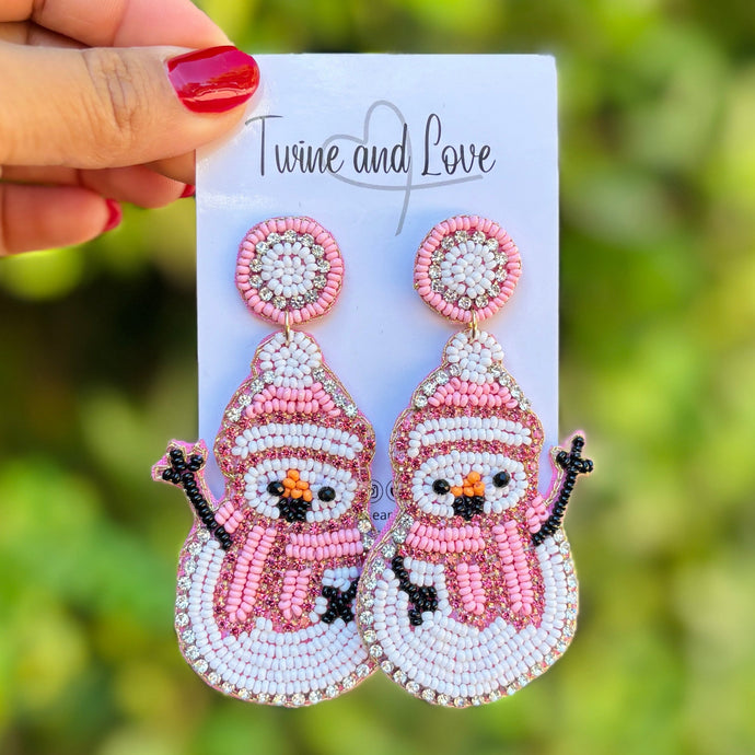 Beaded snowman earrings featuring pink hats and scarves, with black arms and vibrant orange noses. The design conveys a playful, festive tone.
