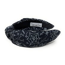 Load image into Gallery viewer, A black fabric headband with a knotted design is patterned with white spiderwebs and spiders. The mood is edgy and Halloween-themed.