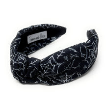 Load image into Gallery viewer, A black fabric headband with a knotted design is patterned with white spiderwebs and spiders. The mood is edgy and Halloween-themed.
