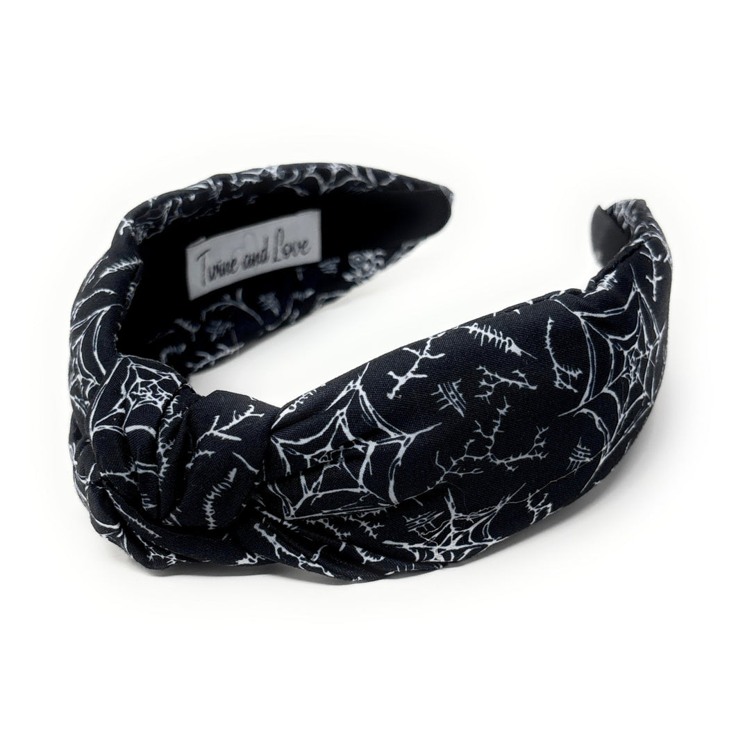 A black fabric headband with a knotted design is patterned with white spiderwebs and spiders. The mood is edgy and Halloween-themed.