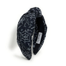 Load image into Gallery viewer, A black fabric headband with a knotted design is patterned with white spiderwebs and spiders. The mood is edgy and Halloween-themed.