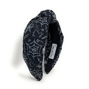 A black fabric headband with a knotted design is patterned with white spiderwebs and spiders. The mood is edgy and Halloween-themed.