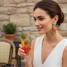 Load image into Gallery viewer, A woman in a white sleeveless dress holds a cocktail glass with an orange drink, mint, and orange slice, wearing a large decorative earring in front of a stone wall background.