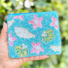 Load image into Gallery viewer, Hand holding a blue beaded pouch with starfish and shell designs against a blurred green background