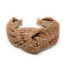 Load image into Gallery viewer, A natural woven raffia or straw twist-front headband with a knotted center and a beige cardboard backing visible at the ends, shown on a plain white background.