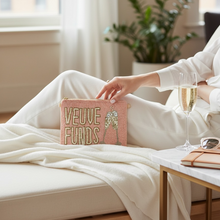 Load image into Gallery viewer, A person in a white robe reclines on a bed holding a pink rectangular clutch or pouch with the words 'VEUVE FUNDS' in gold lettering and a small hanging charm. A champagne flute, sunglasses, and a notebook sit on a bedside table in a softly lit bedroom.