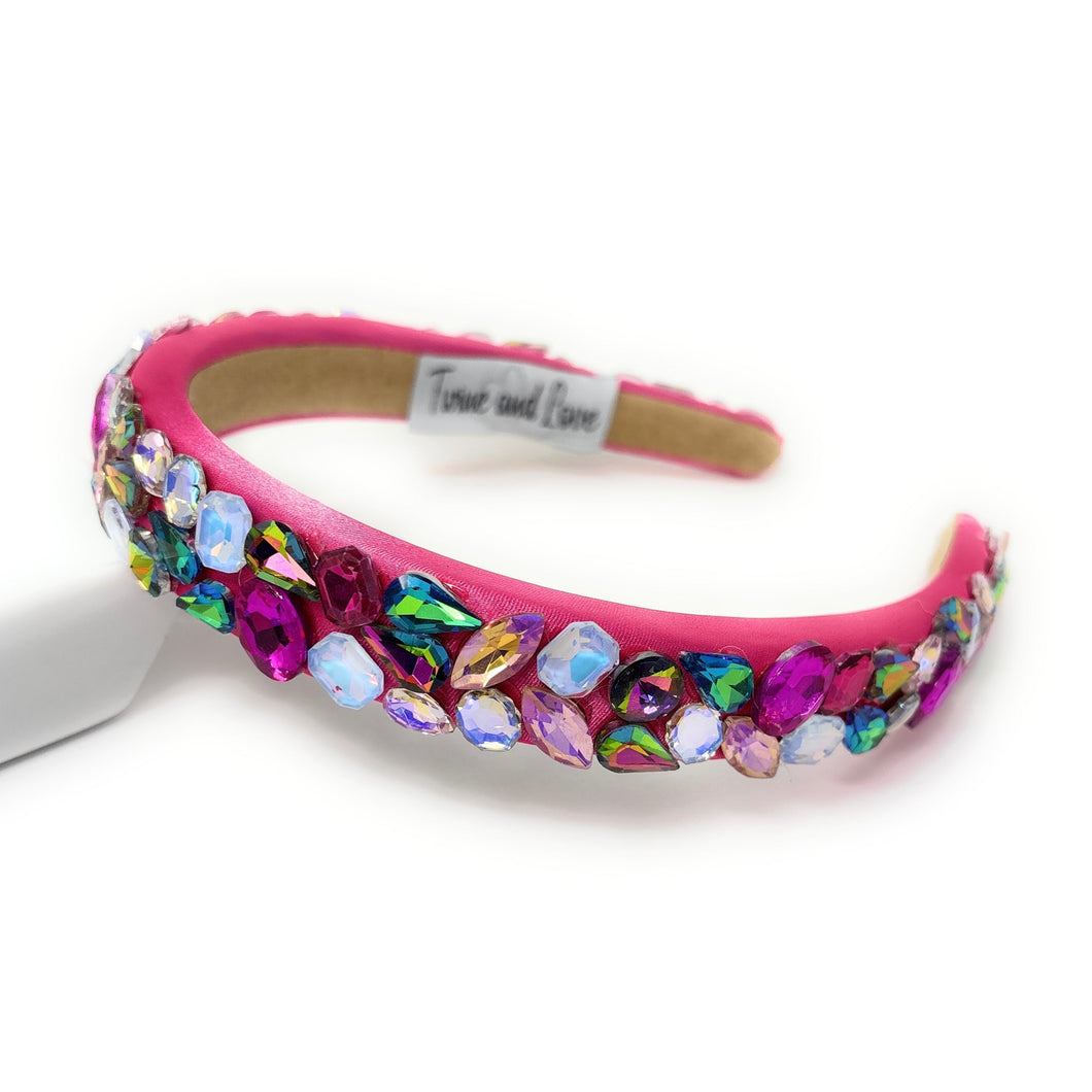 A vibrant pink headband adorned with multicolored gemstones, including blue, purple, and green. It exudes a playful, glamorous vibe.