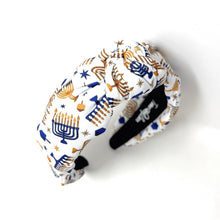 Load image into Gallery viewer, A fabric headband with a white background and a festive pattern of gold and blue menorahs, stars, and candles, shown on a plain white background.