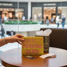 Load image into Gallery viewer, A hand holds a yellow woven clutch bag with pink text reading 'I'M BUYING AGAIN' on a round wooden table in a modern indoor setting. A patterned tissue box and a small card sit beside it, with a blurred mall interior in the background.