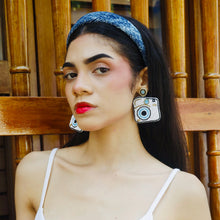Load image into Gallery viewer, A chic model with a unique style, wearing a white outfit and oversized camera earrings against a wooden backdrop.
