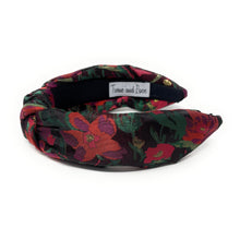 Load image into Gallery viewer, A colorful, padded headband with a floral pattern in red, green, and black hues. The fabric is twisted at the front, creating a knot-like design.