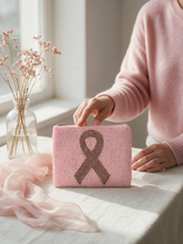 Load image into Gallery viewer, A pink pouch with a pink ribbon design sits on a white tablecloth, with a person in a pink sweater reaching toward it. A glass vase with dried flowers and a sheer pink scarf are nearby.