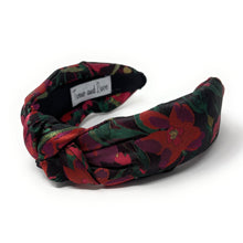 Load image into Gallery viewer, A colorful, padded headband with a floral pattern in red, green, and black hues. The fabric is twisted at the front, creating a knot-like design.
