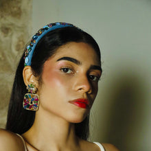 Load image into Gallery viewer, A fashionable woman wearing a blue headband and colorful earrings, set against a blurred background of bottles.
