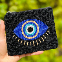 Load image into Gallery viewer, A hand holding a black coin purse adorned with a beaded blue eye design and a gold outline, with a blurred background.